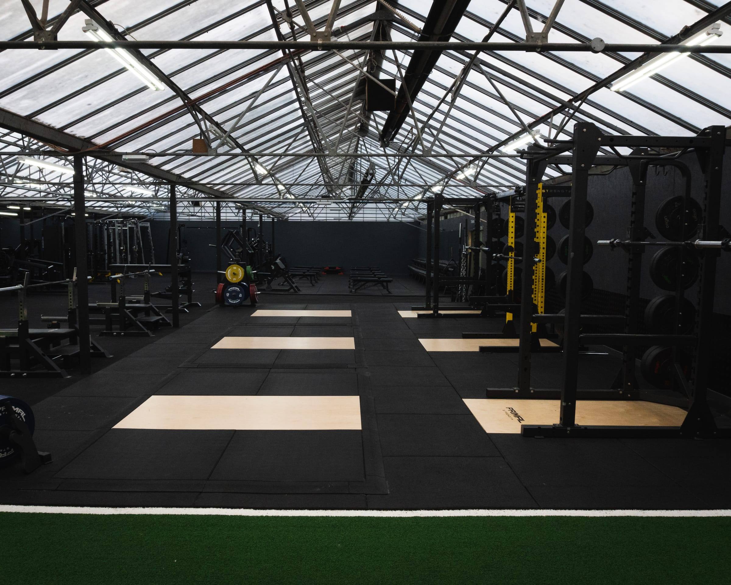 Base Gym Bristol: lifting platforms and power racks beneath the glazed roof, with the turf sled track in the foreground